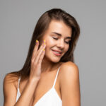 Portrait of beautiful young brunette with bare shoulders touching her cheek, looking at camera and smiling, on gray background